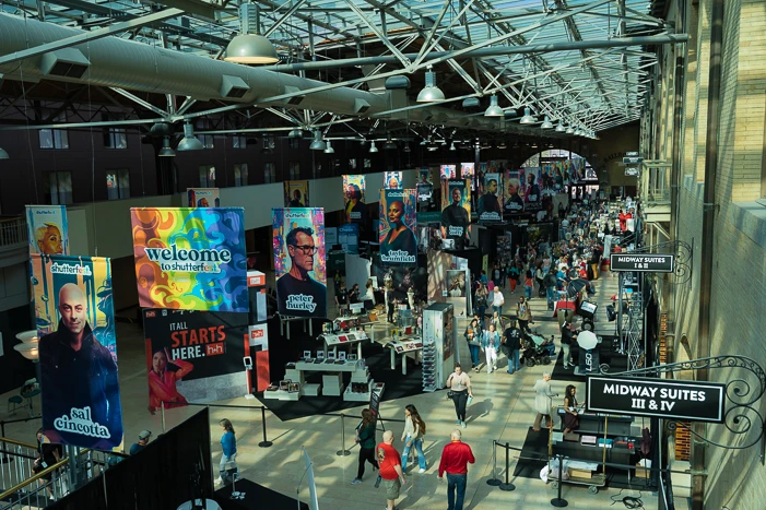 Overhead view of a large indoor convention or trade show hall with crowds of people walking between vendor booths and displays.