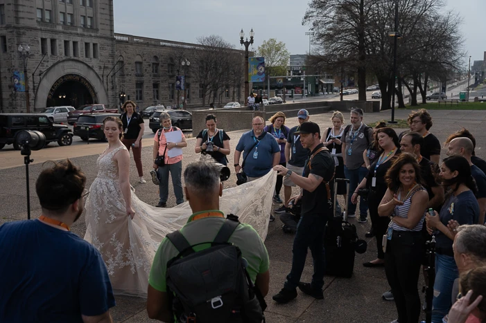 Bride in white dress holding veil fabric surrounded by photographers and crew on urban street.