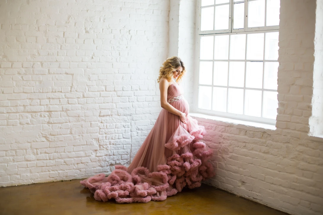 Pregnant woman in pink tulle gown gazing toward large industrial window with white brick walls.