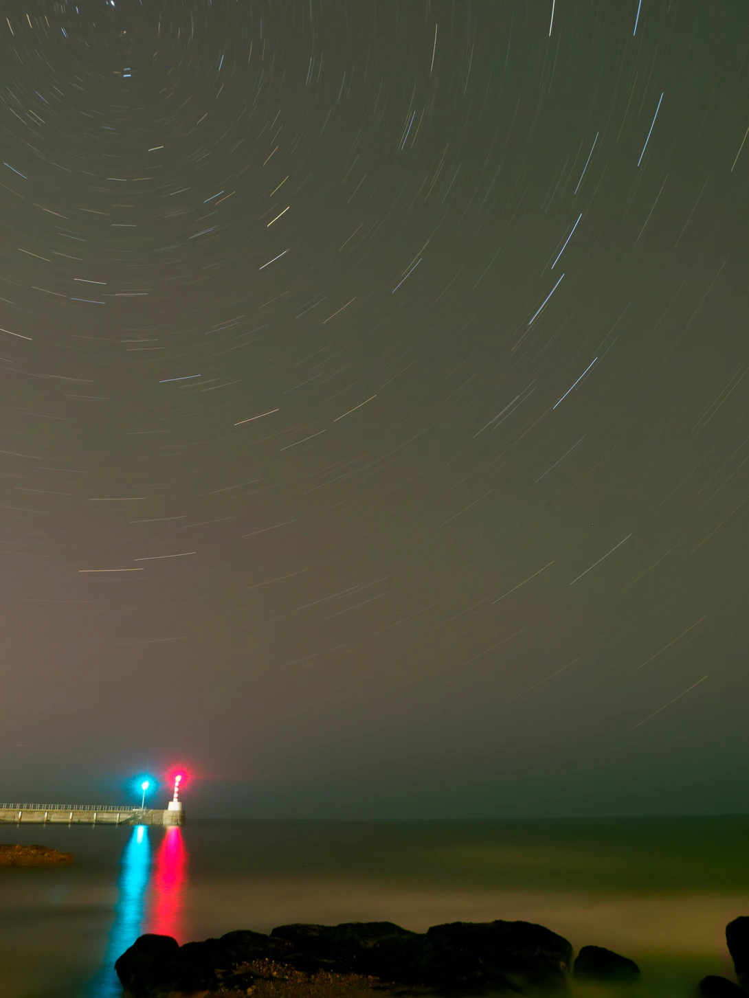 Night sky long exposure with star trails above silhouetted landscape and colorful light trails in foreground.