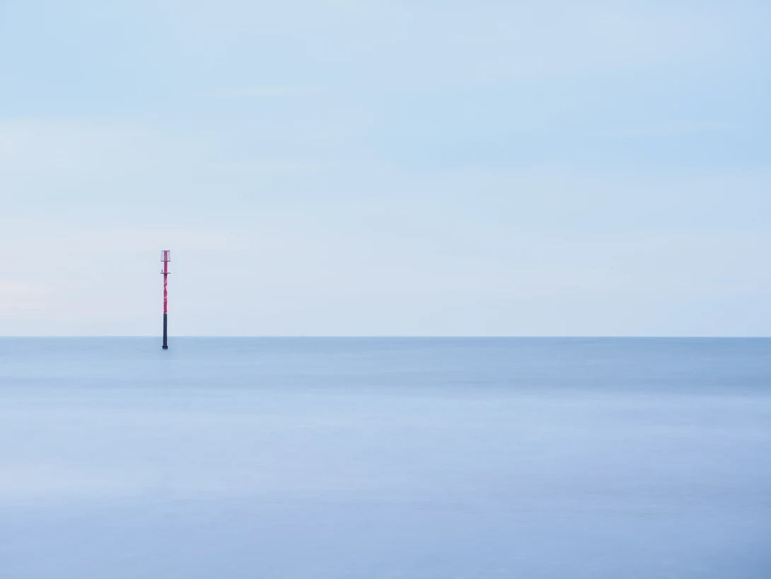 A single vertical pole or marker in calm shallow water under a clear blue sky.