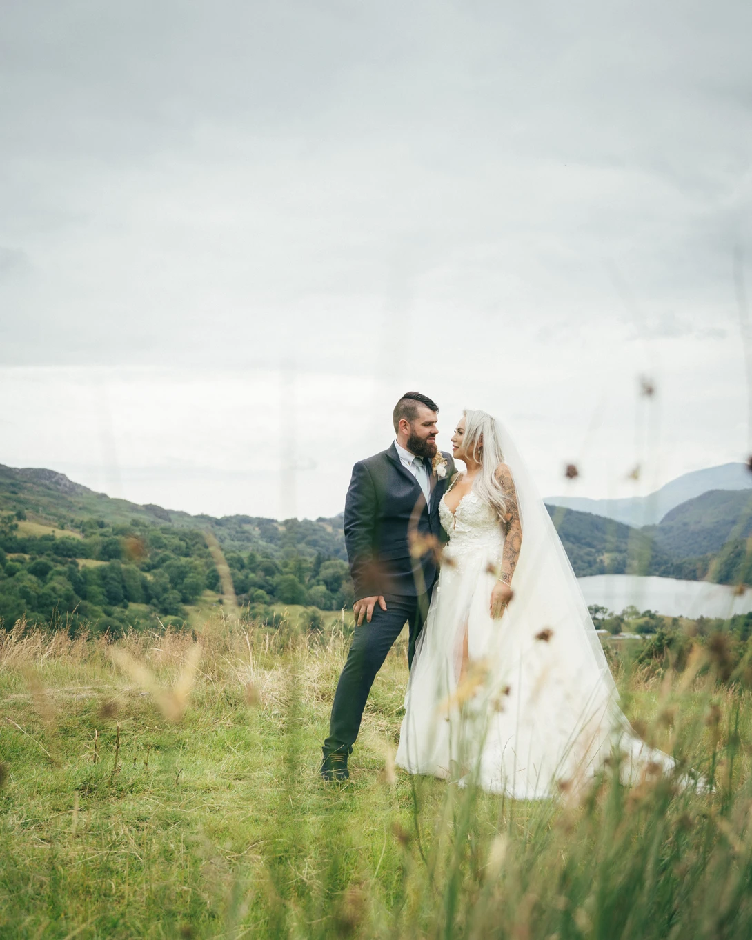 Bride and groom standing together in a grassy field with mountains and water visible in the background.