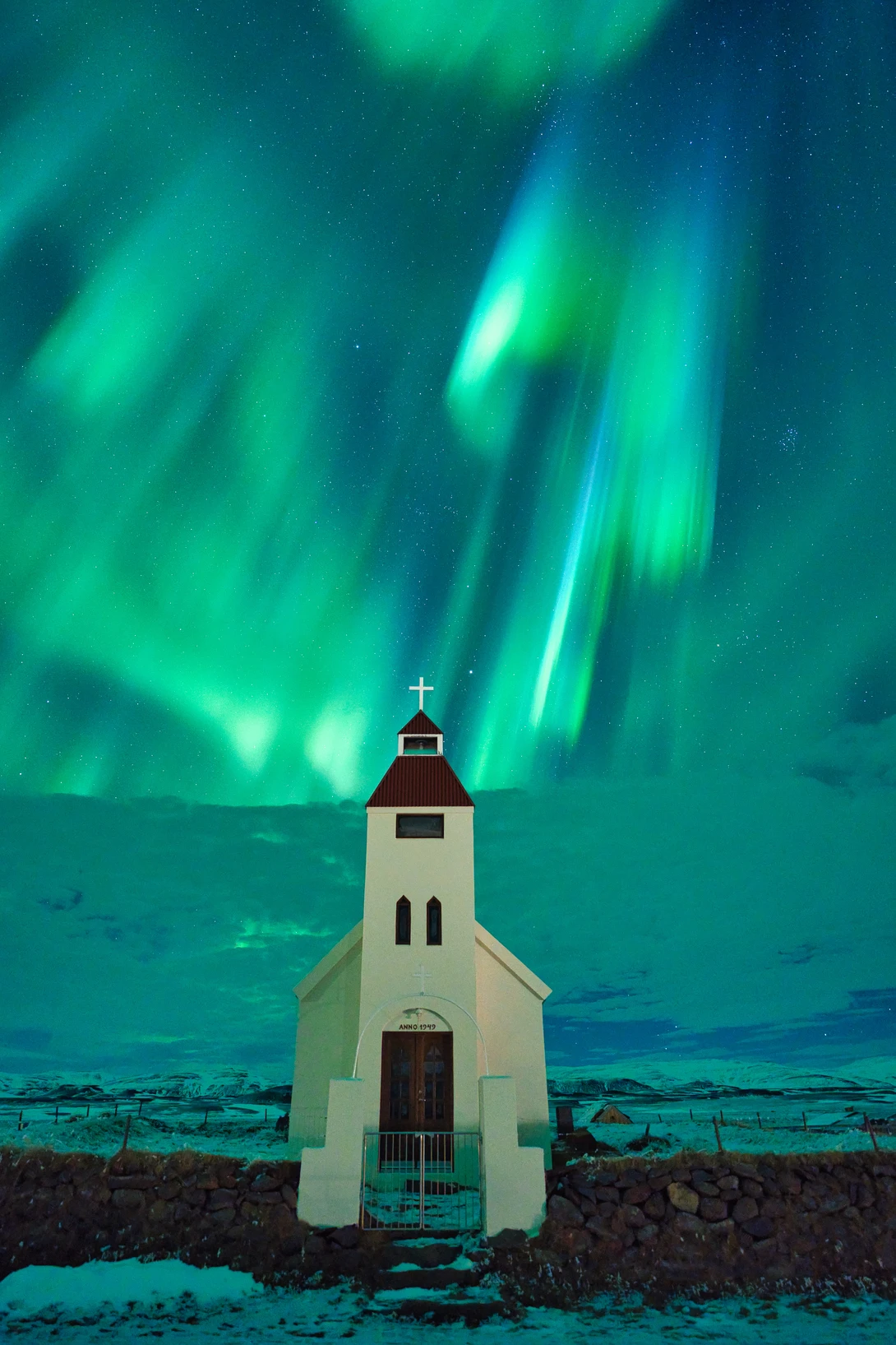 White wooden church with steeple silhouetted against vibrant green aurora borealis dancing across night sky.