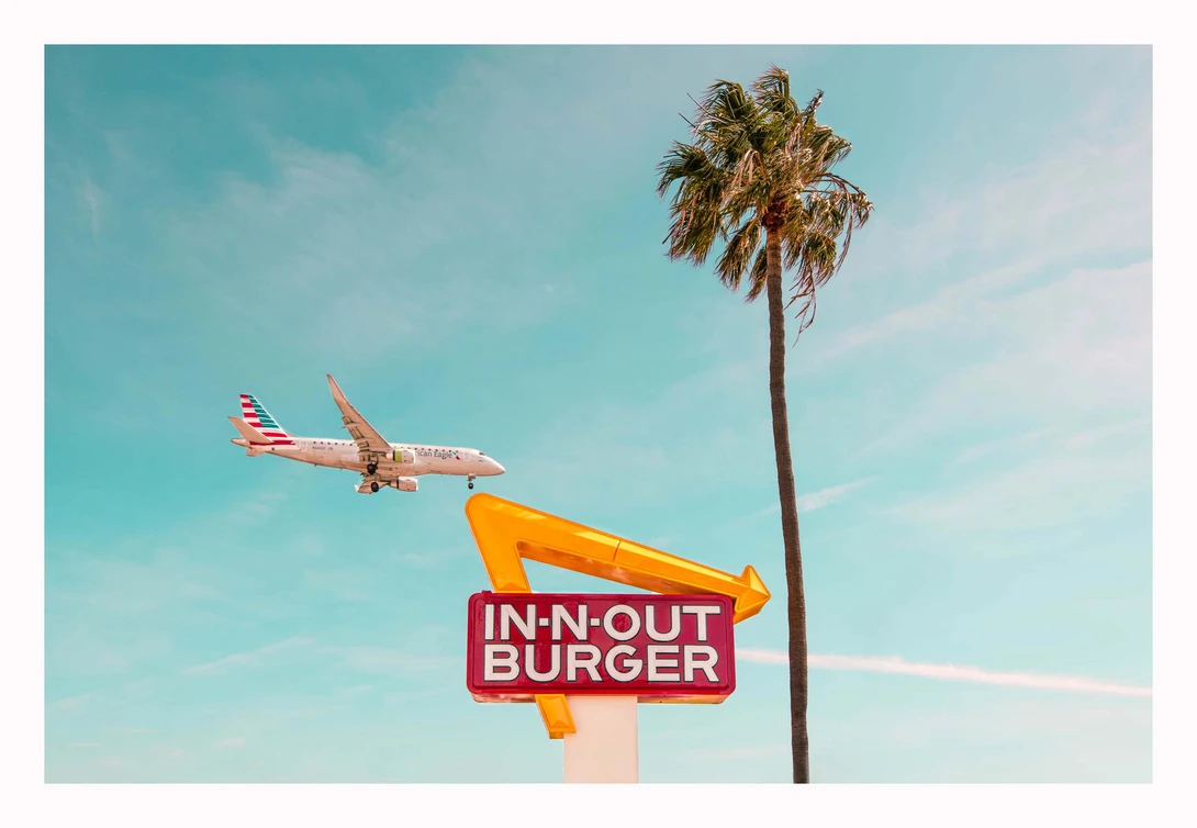 Airplane flying over an In-N-Out Burger sign with a palm tree against a blue sky.