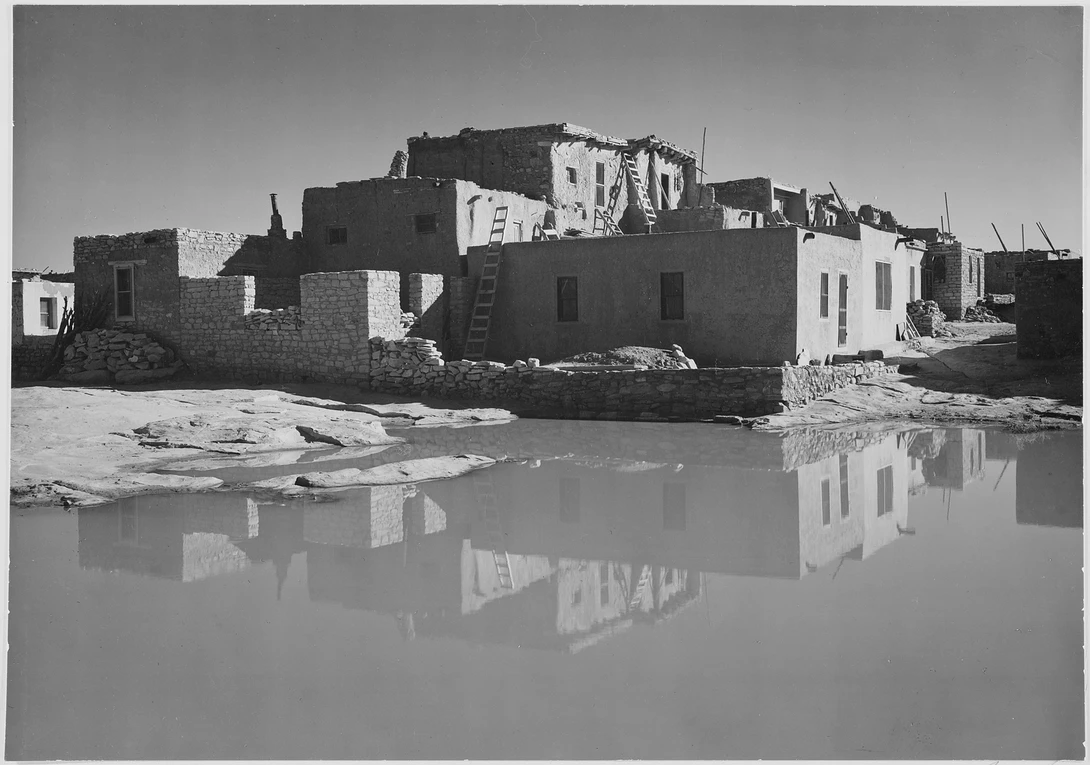 Black and white photograph of deteriorating adobe buildings reflected in still water.