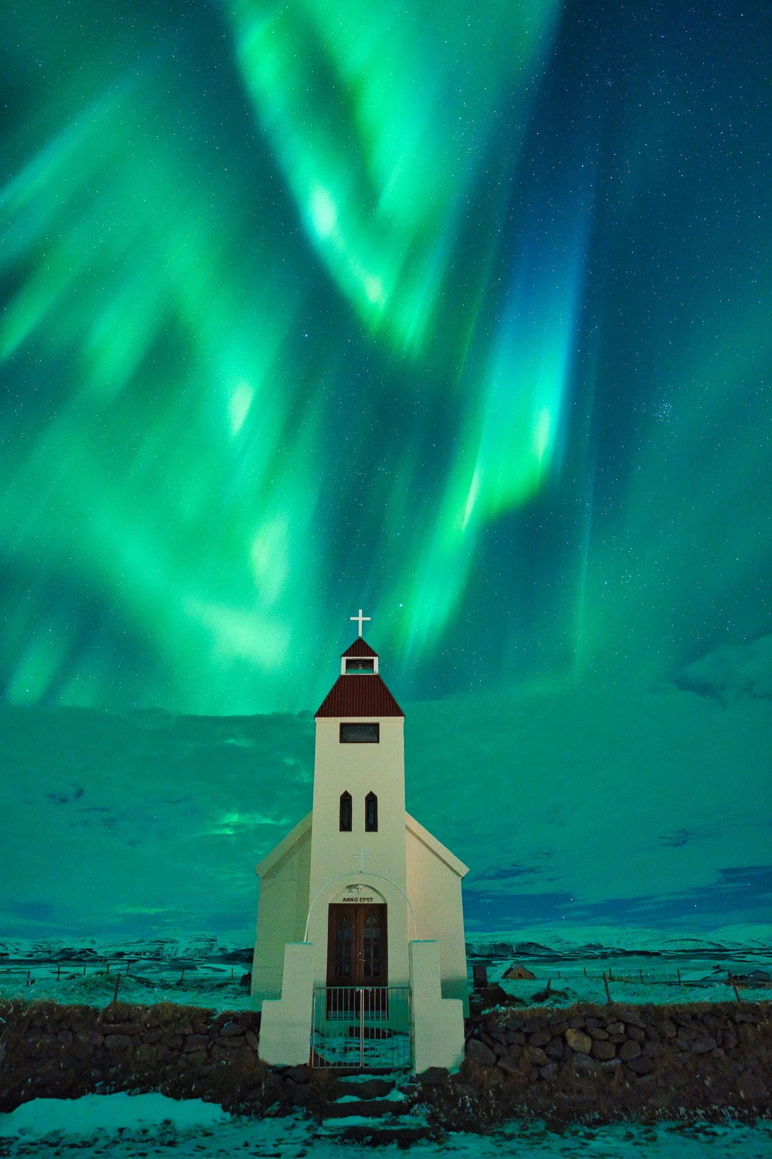 Small white church with cross silhouetted against vibrant green aurora borealis over dark ocean.