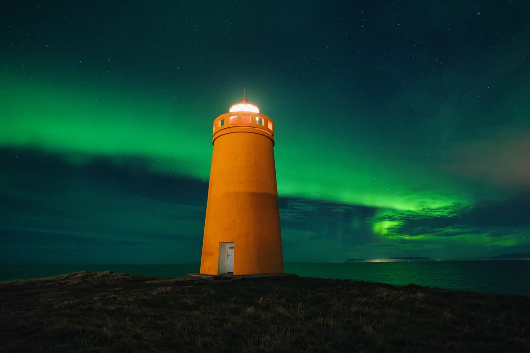 Orange lighthouse illuminated against a vibrant green aurora borealis in a dark night sky.
