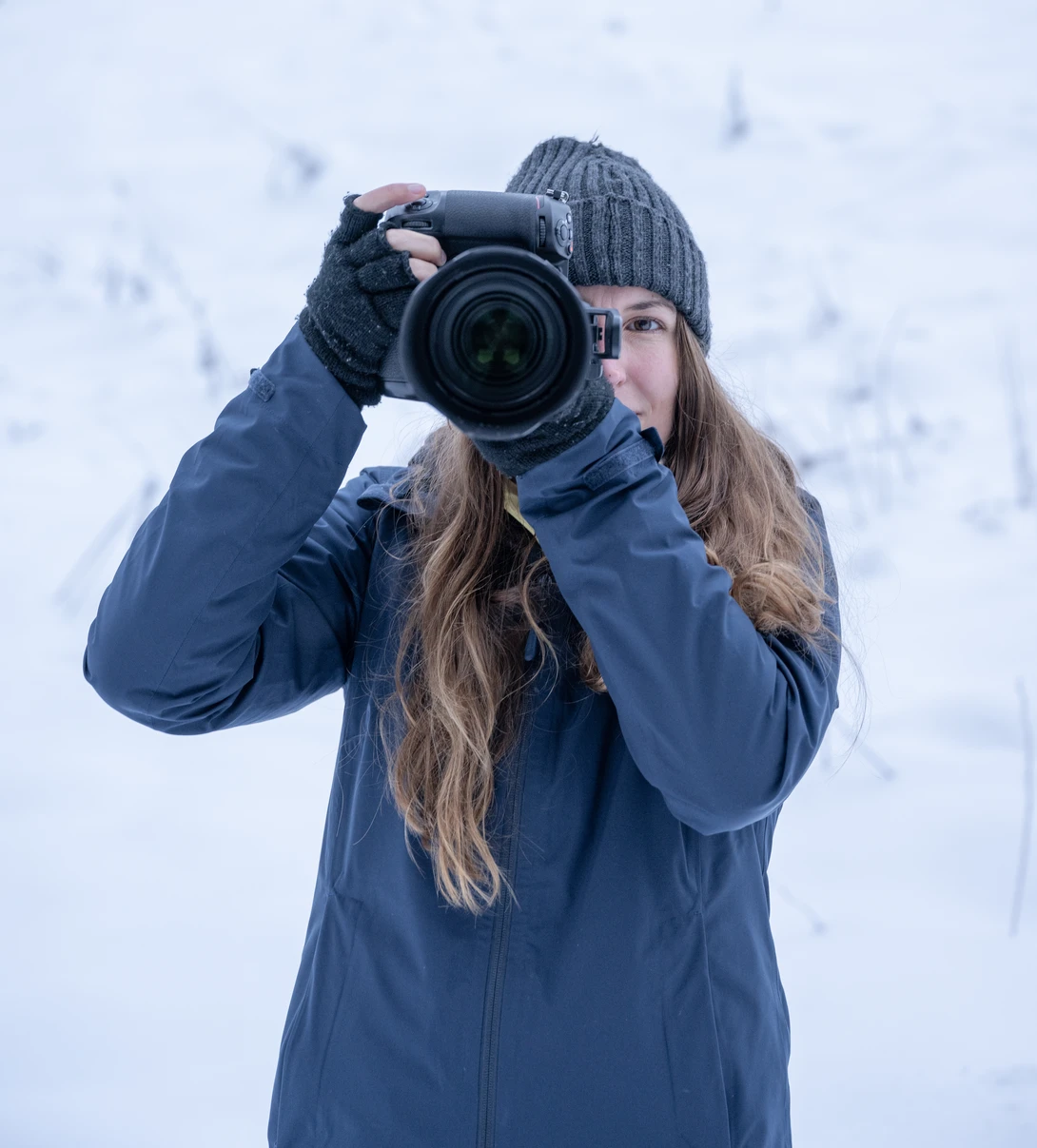 Photographer holding a DSLR camera up to her face in snowy conditions.
