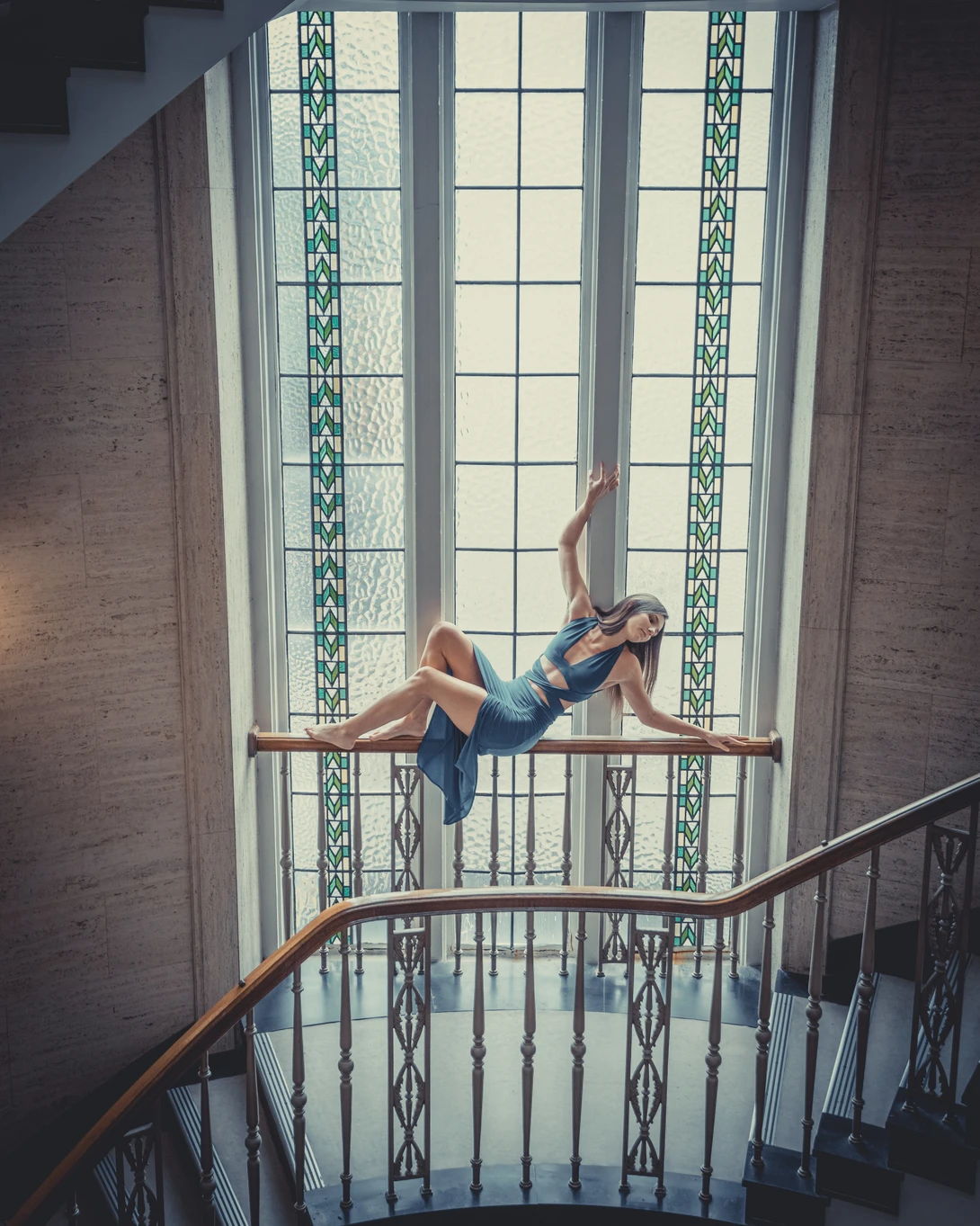 Woman in casual clothing reclines on a metal railing within a stairwell, framed by a large window with geometric tile patterns.