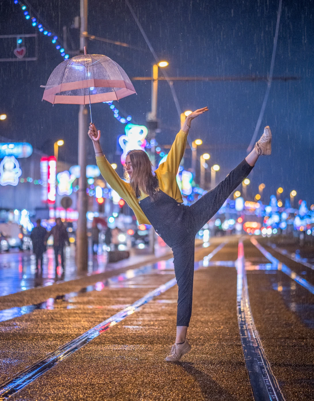 Dancer performing a high kick on a wet city street at night, holding a transparent umbrella.