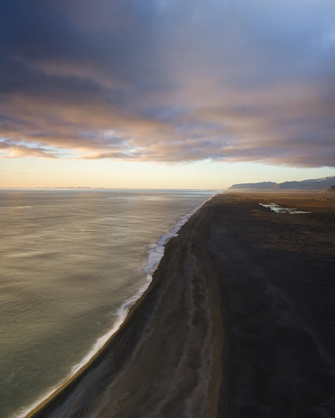 Dramatic coastal cliff with black sand stretching into the ocean under a golden-hour sky.