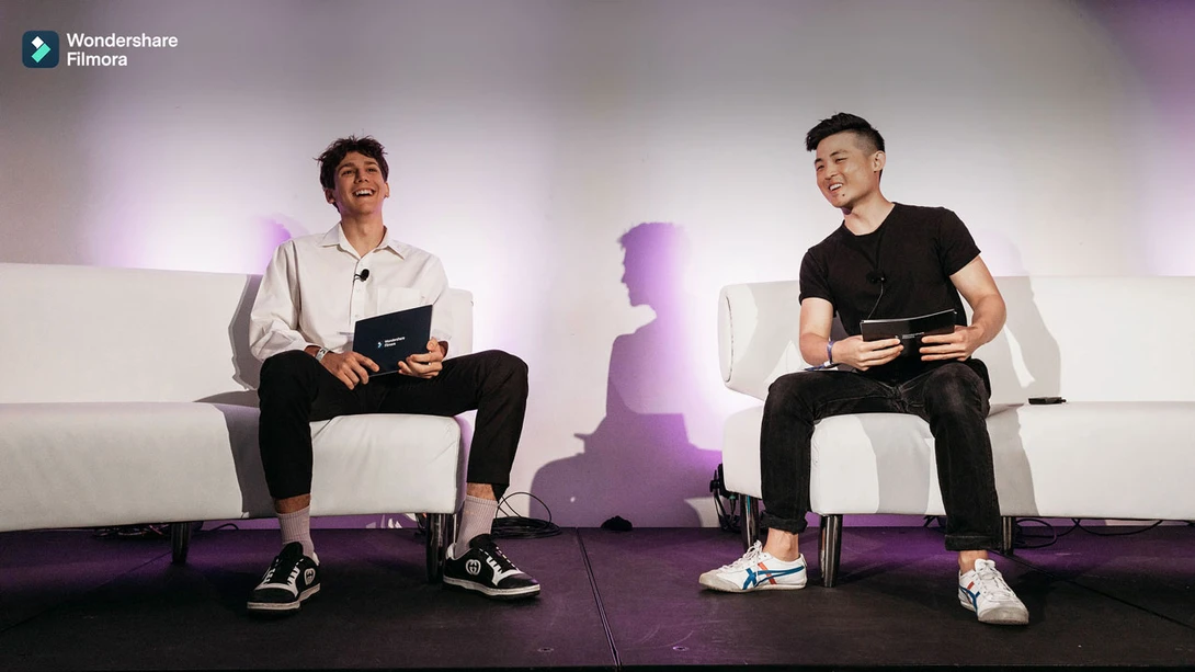 Two men seated on white chairs during a panel discussion with purple stage lighting and backdrop.
