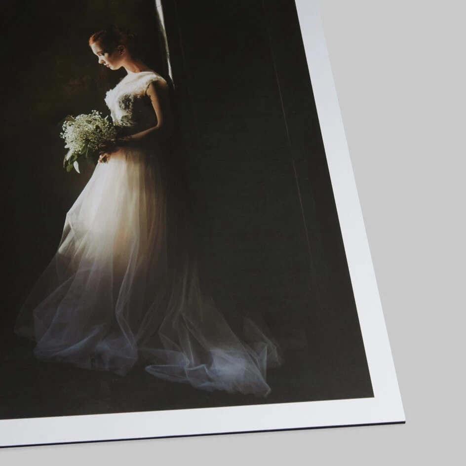 Bride in flowing white gown holding bouquet against dark draped backdrop, dramatic side lighting.