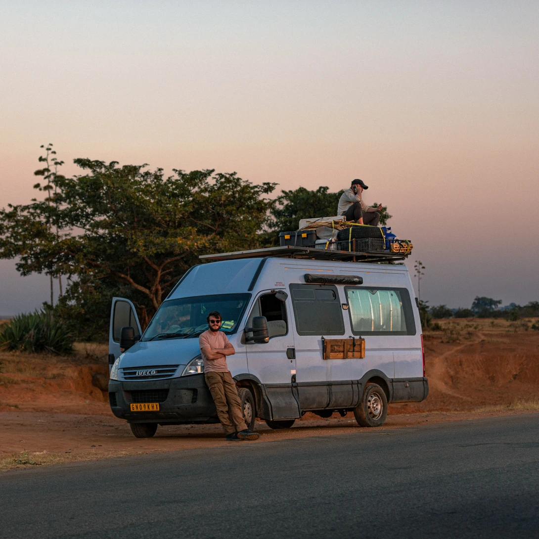 Camper van parked on a roadside at dusk with travelers on the roof rack and a large tree nearby.