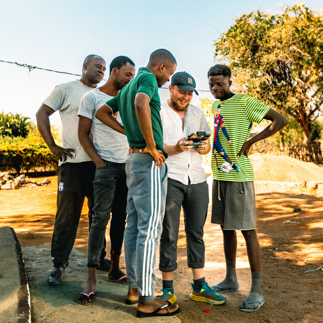 Six people gathered around a smartphone in an outdoor rural setting during golden hour.