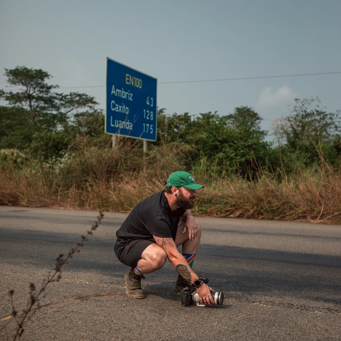 Man crouching on a road beneath a blue directional sign in a rural area.