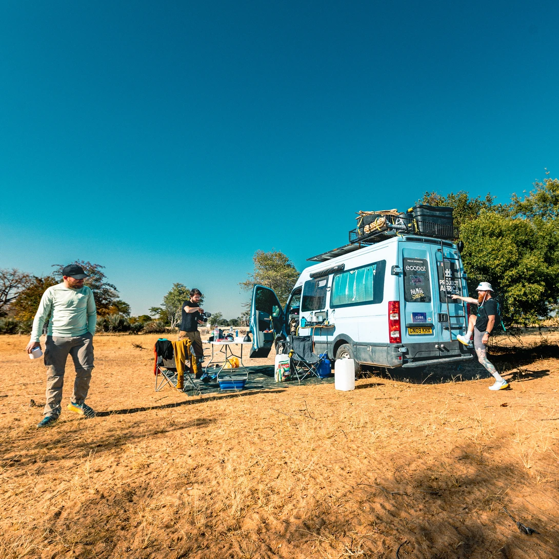 A white van parked in a dusty field with people and equipment nearby under clear blue sky.