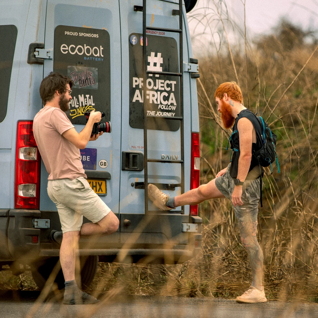 Two people outside a converted van with project signage, one adjusting equipment near the vehicle door.