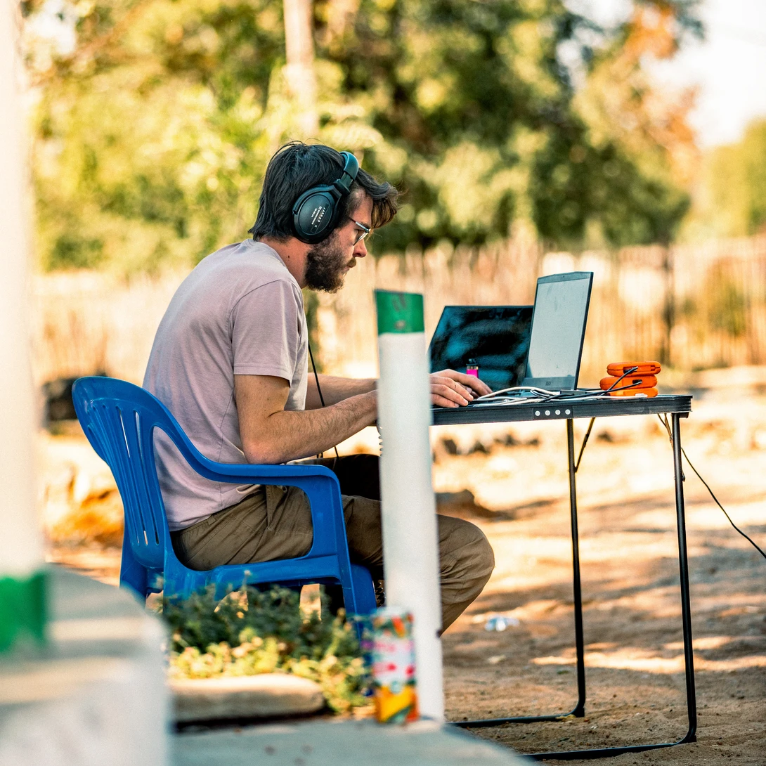 Freelancer working outdoors at a folding table with laptop and coffee, wearing headphones and casual clothing.