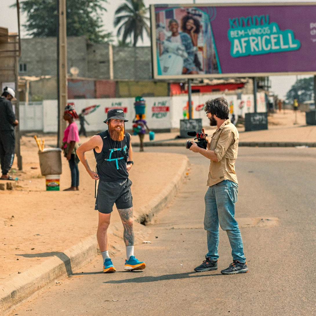 Two people stand in an African street under bright sunlight, with buildings and a large billboard visible behind them.