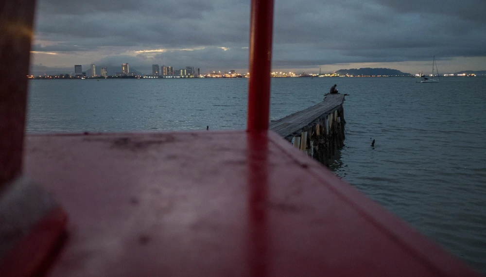 Wooden pier extending into calm water at dusk, with a city skyline glowing in the distance across the bay.