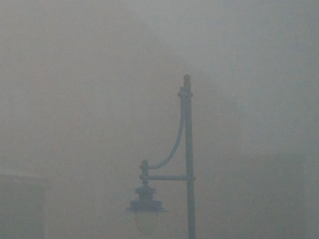 Ornate street lamp silhouetted against heavy fog obscuring urban buildings in background.