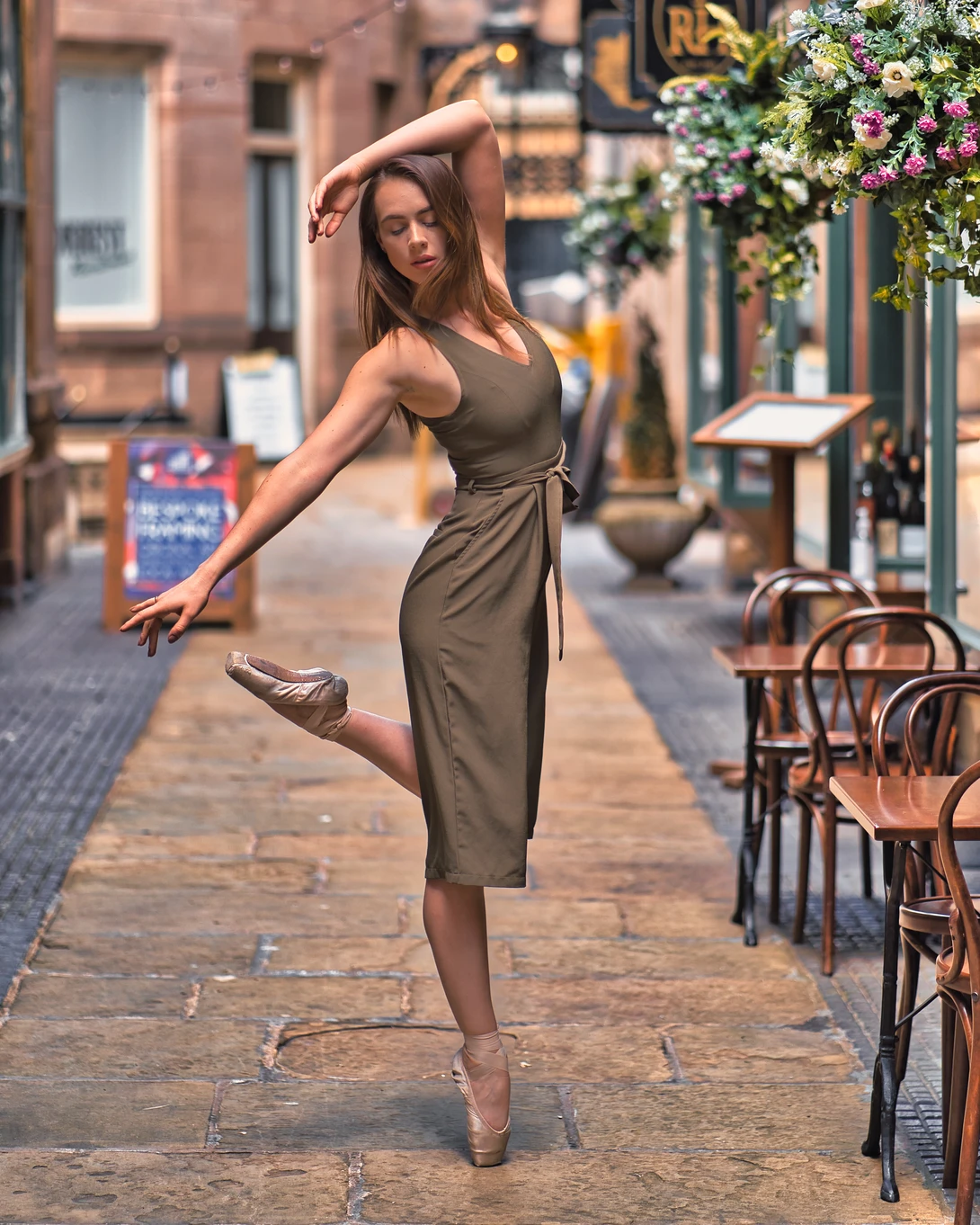 Dancer in olive dress performing en pointe on a European cobblestone street lined with brick buildings and cafe chairs.