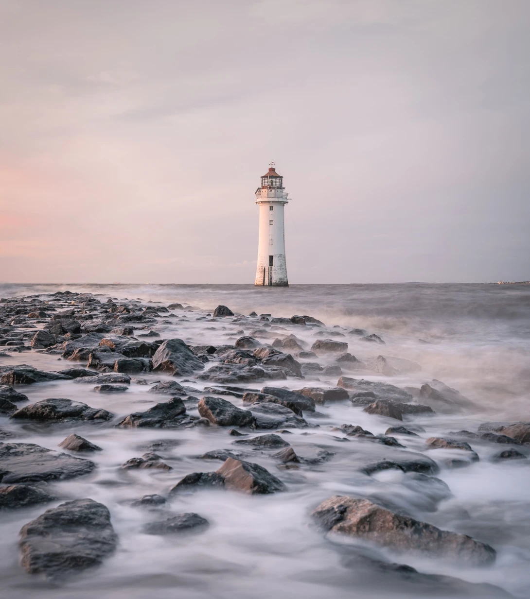 White lighthouse standing in shallow water among dark rocky foreground during golden hour.