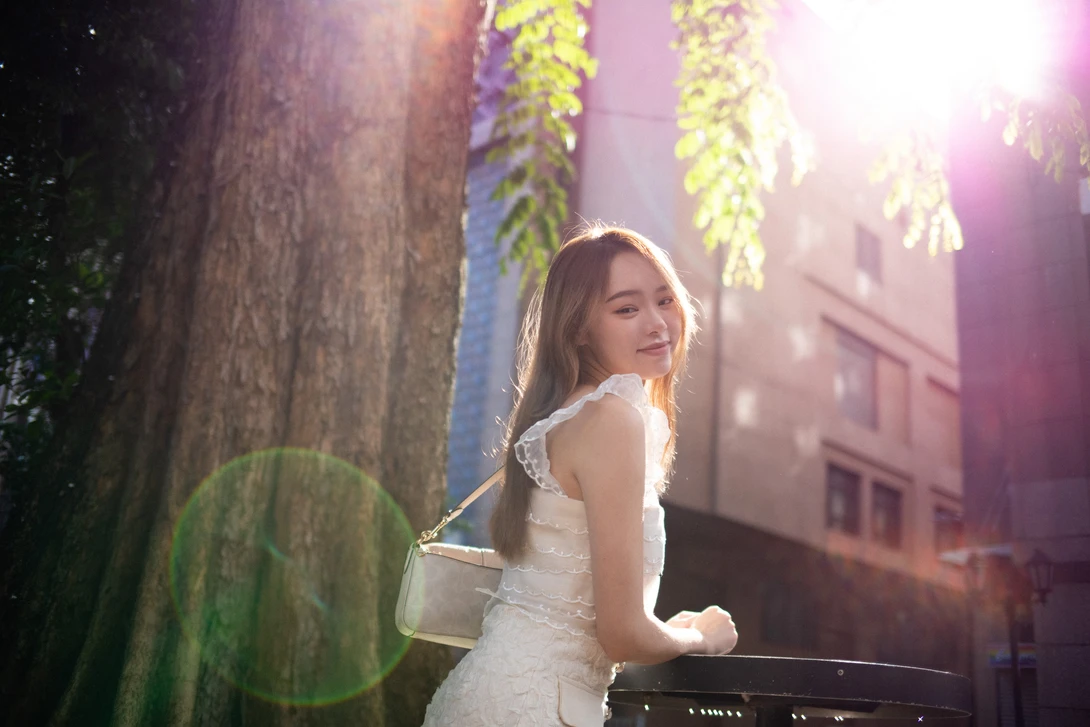 Woman in white dress looking over shoulder in golden sunlit setting with large tree and building backdrop.