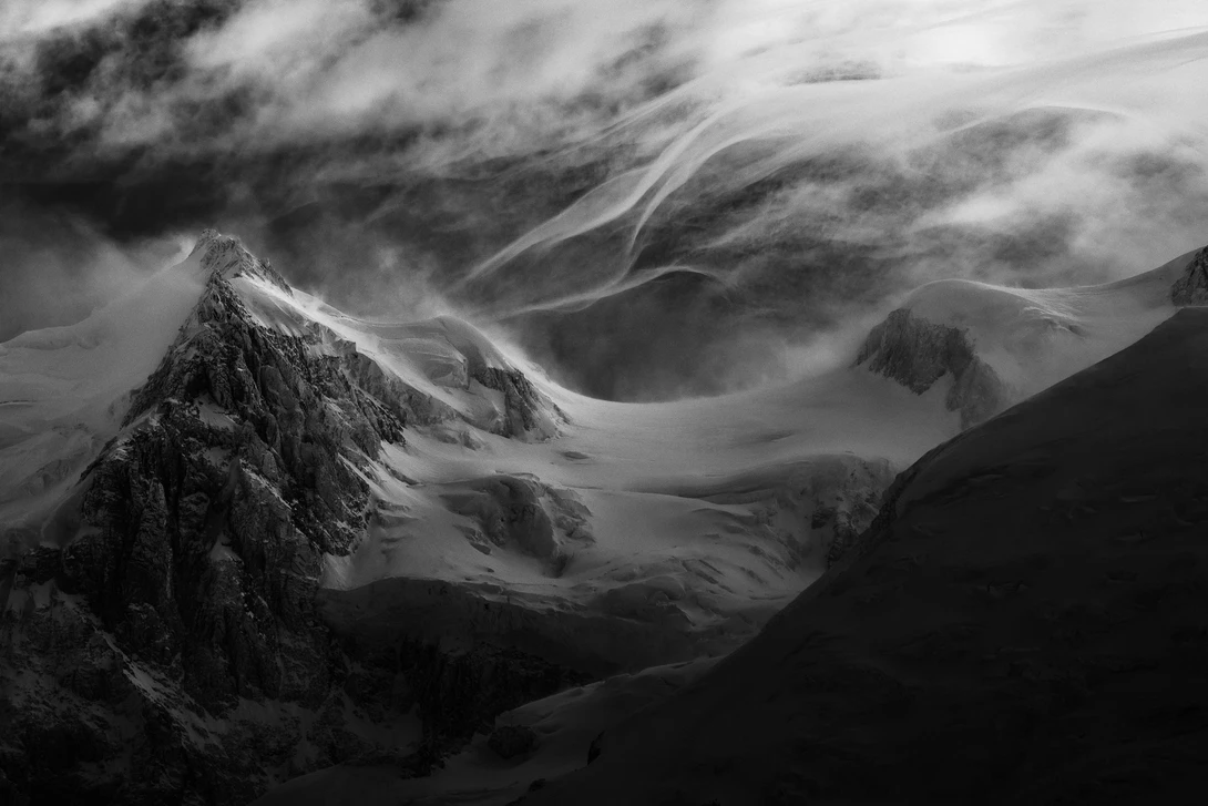 Snow-covered mountain peaks and glacier valleys beneath dramatic storm clouds in black and white.
