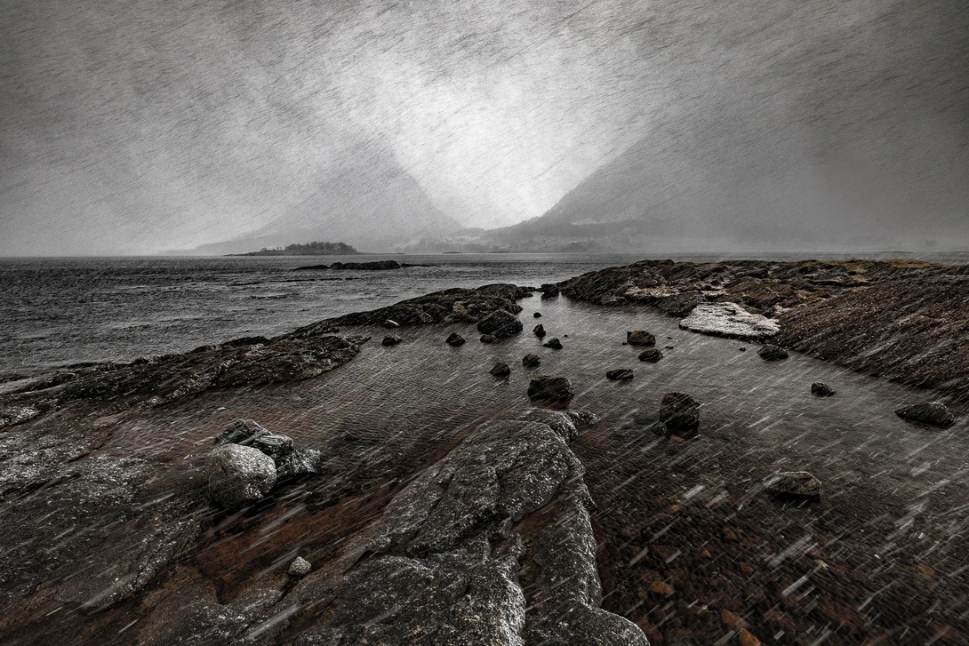 Rocky shoreline with scattered boulders beneath dramatic storm clouds and mountainous landscape.