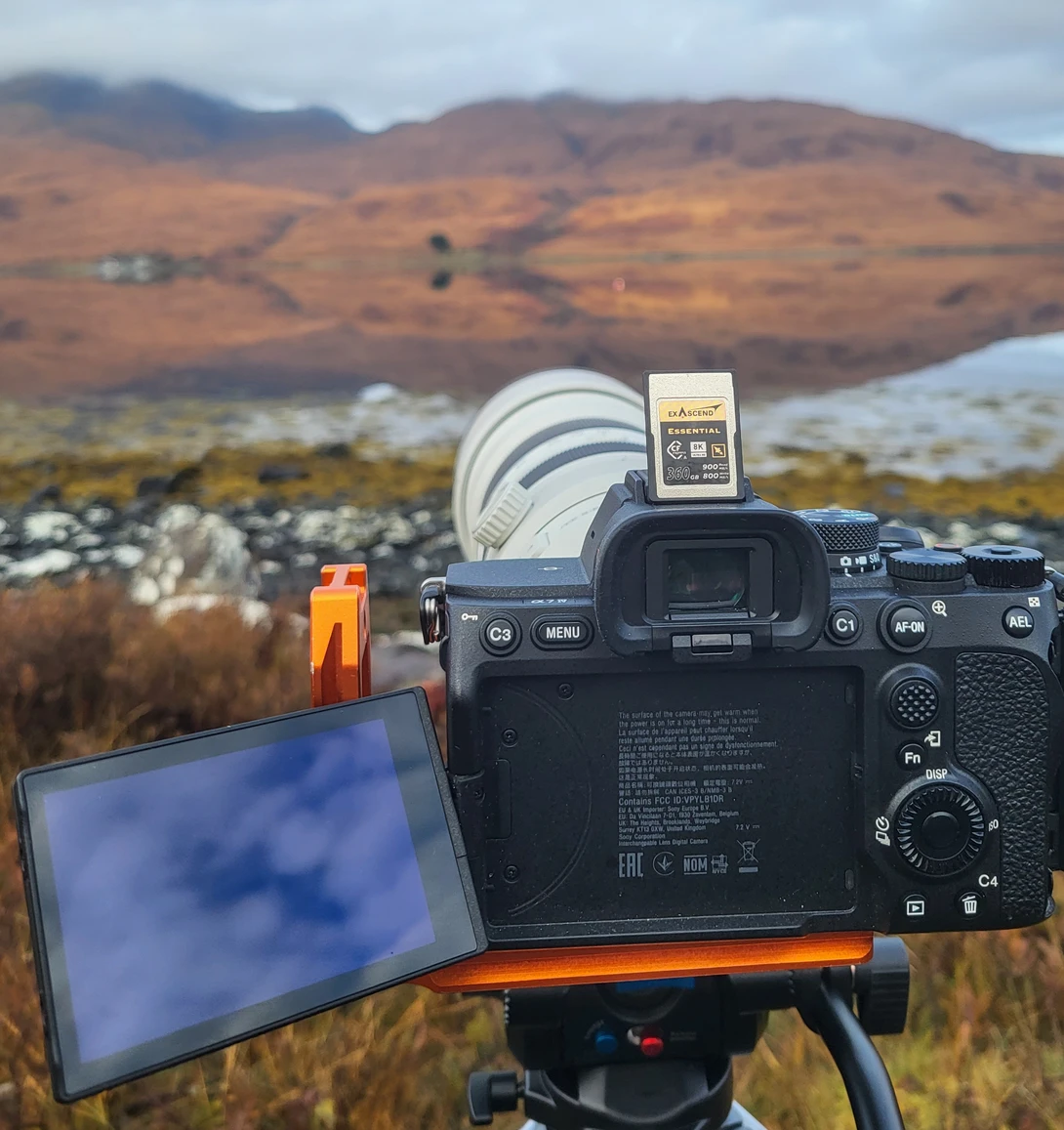 Camera mounted on tripod with articulating monitor displaying live view, mountainous landscape in background.
