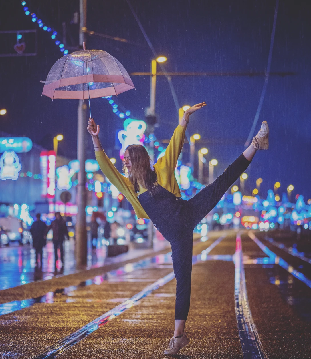 Dancer with arms raised holding a transparent umbrella on a wet urban street at night.