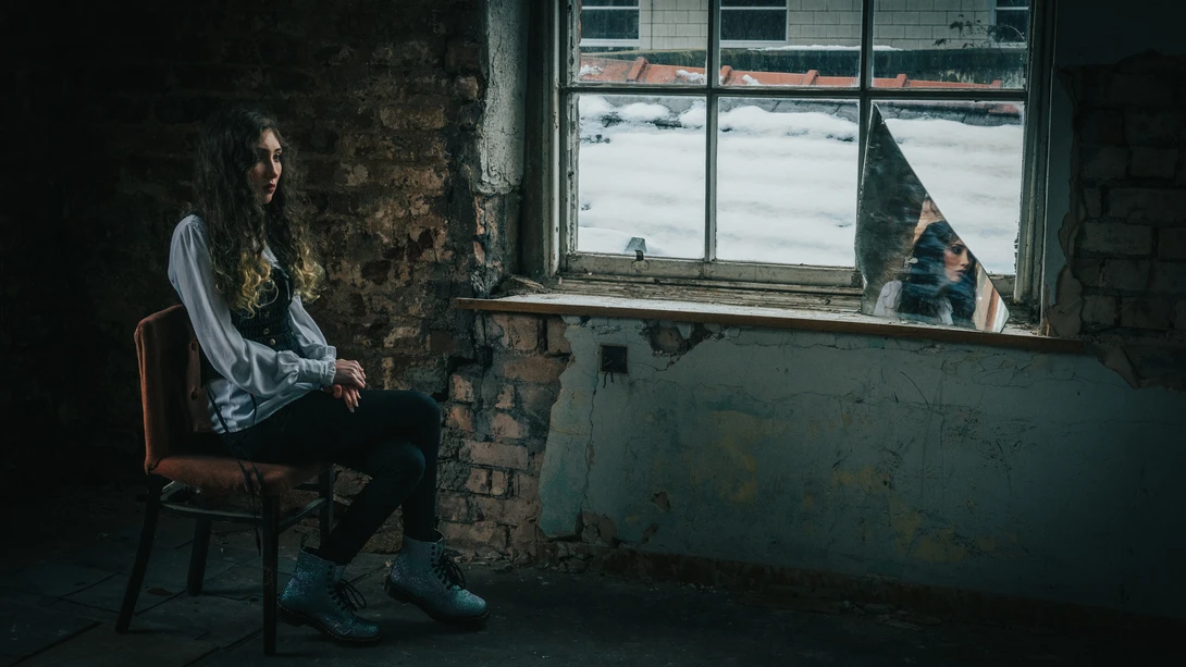Person seated on a wooden chair beside a deteriorating brick wall, gazing out through a weathered window frame.