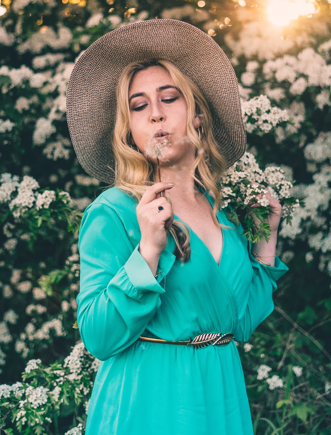 Woman in turquoise dress and wide-brimmed hat posing among white flowering shrubs during golden hour.