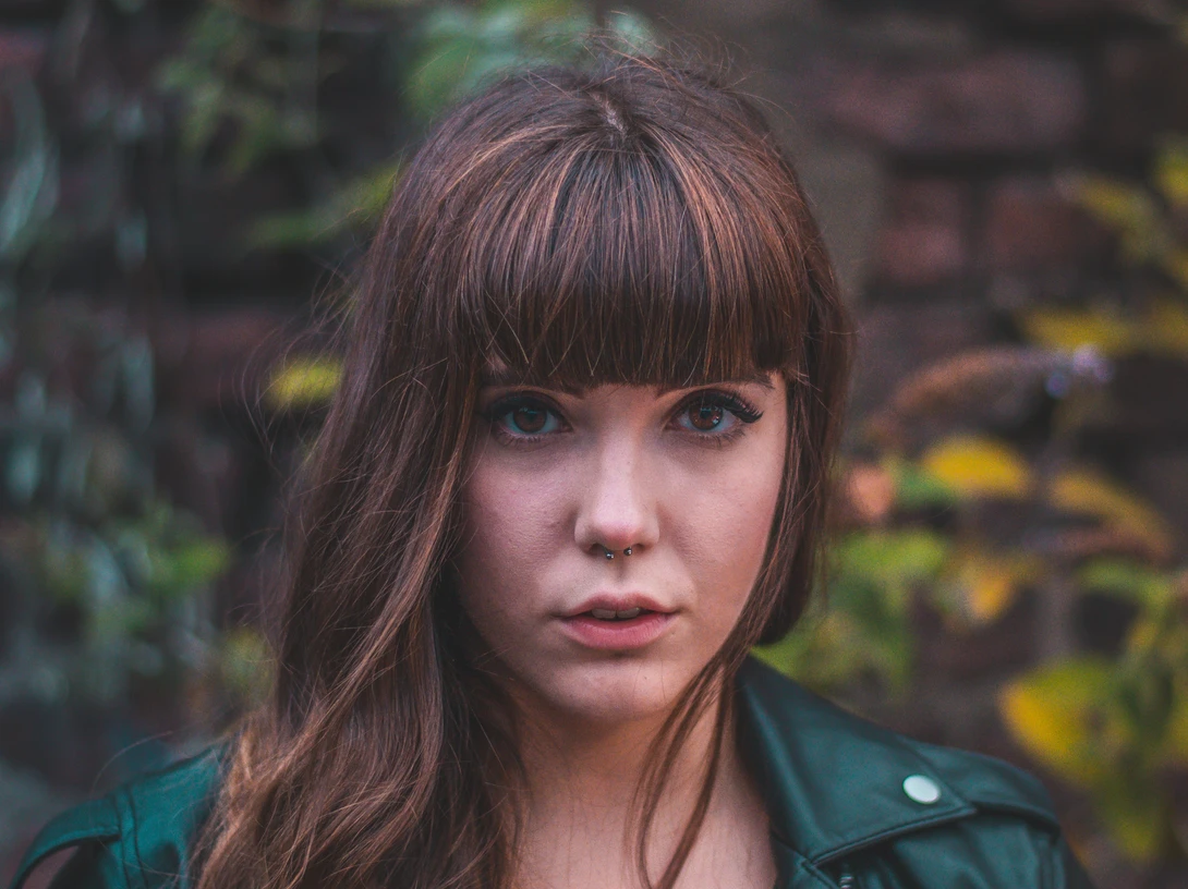 Woman with long brown hair and bangs looking directly at camera against blurred garden background.