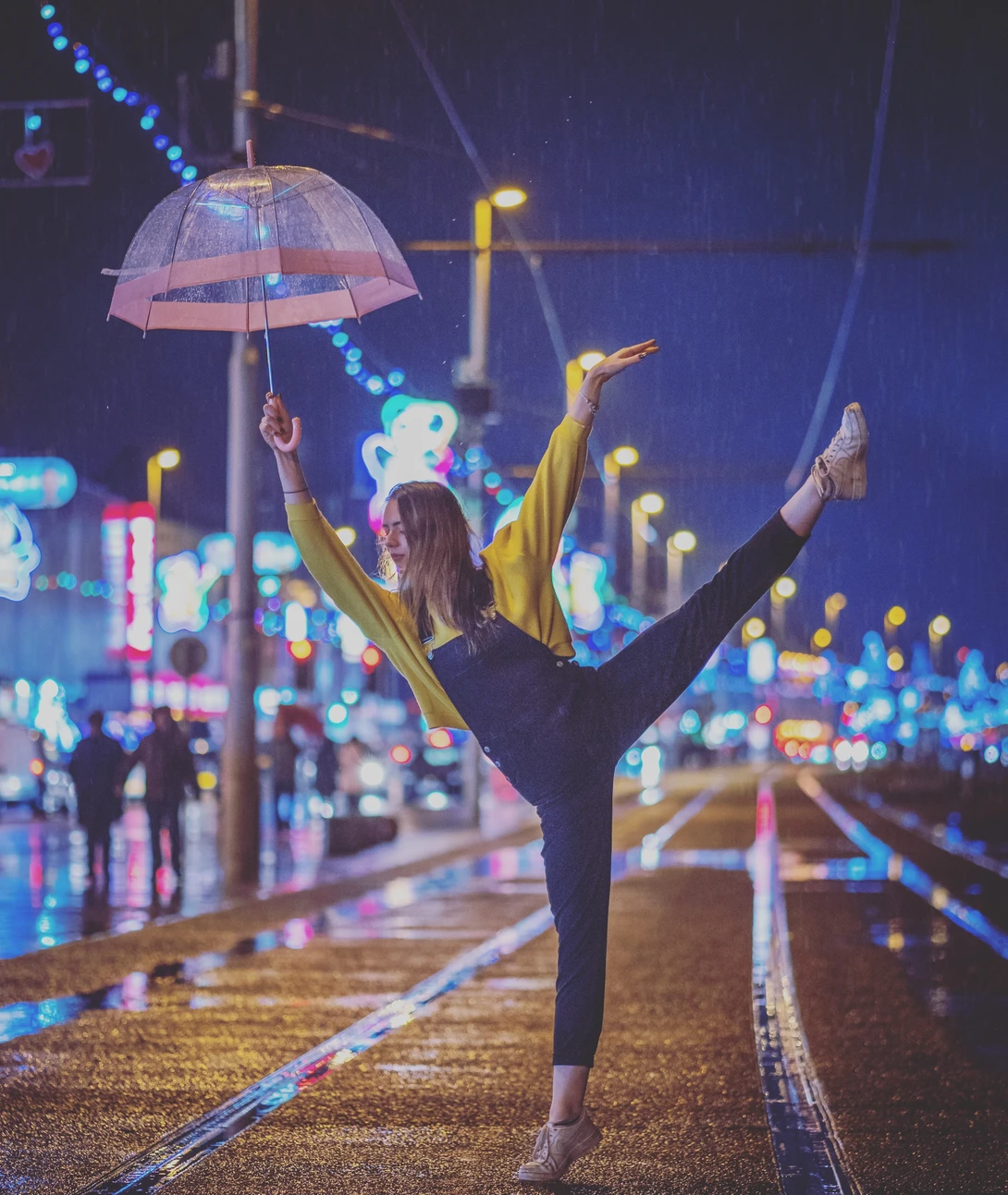 Dancer in black clothing holding a transparent umbrella with arms raised on a wet city street at night.