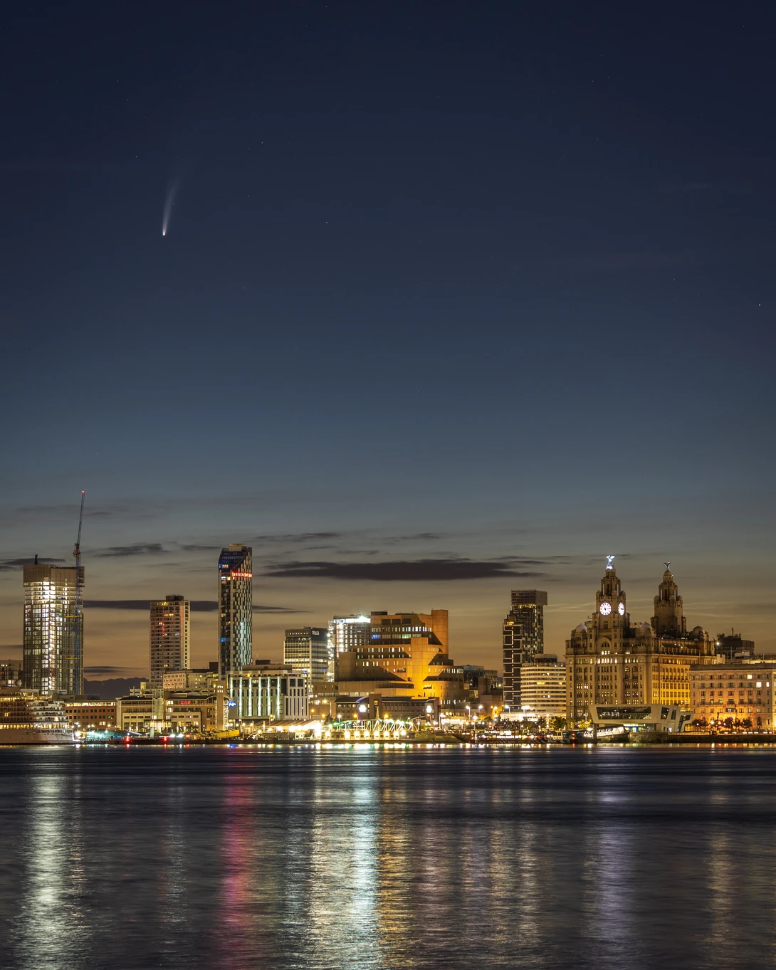 Illuminated waterfront skyline at twilight with reflections in calm water below.