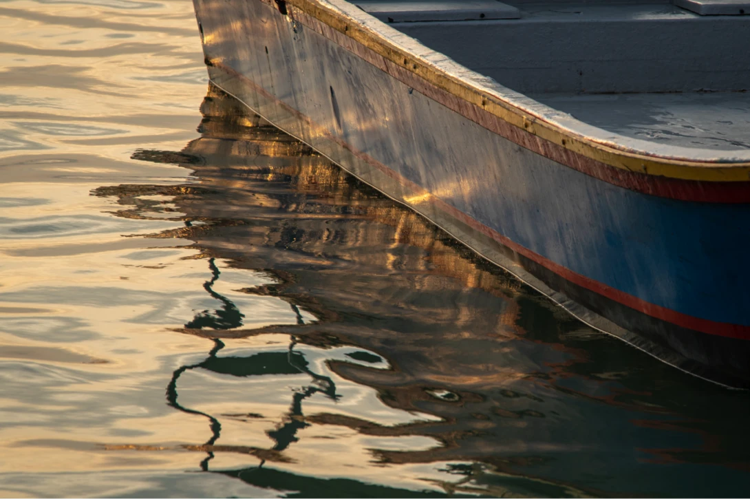 Weathered boat hull reflected in calm golden-hour water with sharp shadow detail.