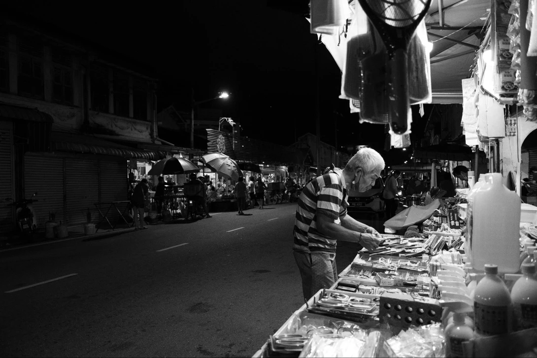 Elderly vendor arranging merchandise at a nighttime street market stall.