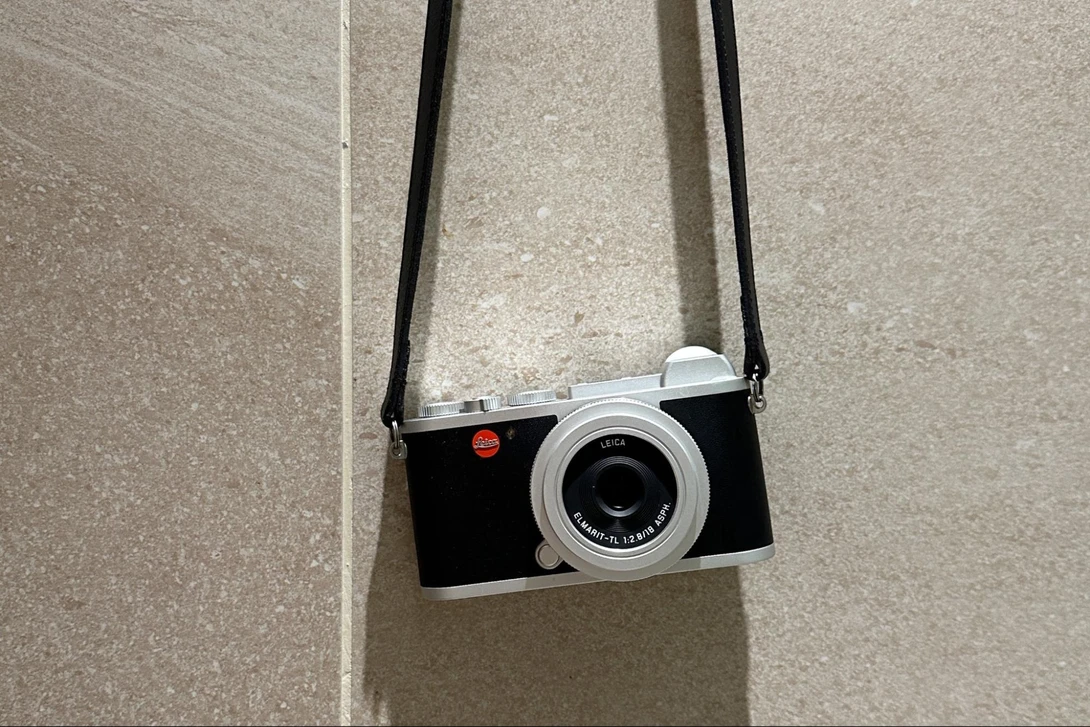 Leica rangefinder camera with black body, silver lens, and red dot logo, suspended by black shoulder strap against concrete wall.