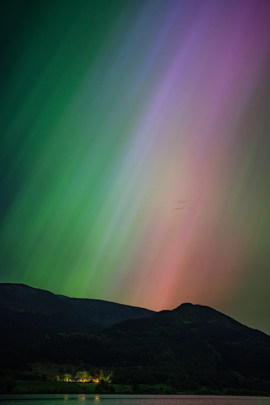 Aurora borealis displaying green and pink bands above a dark mountain silhouette and illuminated shoreline.