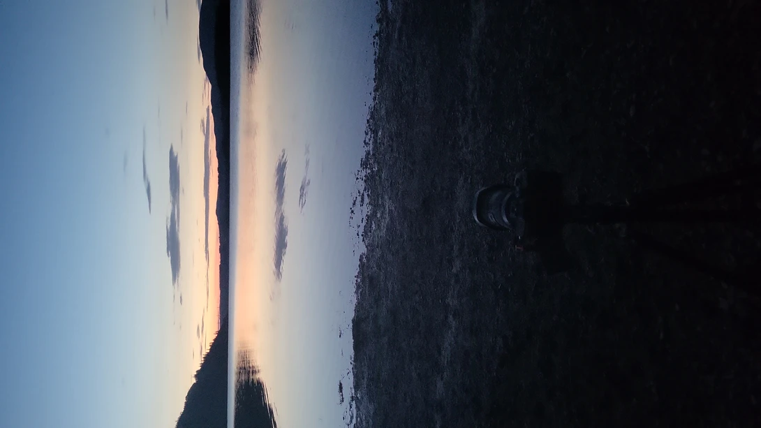 Camera lens in foreground pointing toward a still lake at dusk with mirrored sky reflections and forested hills.