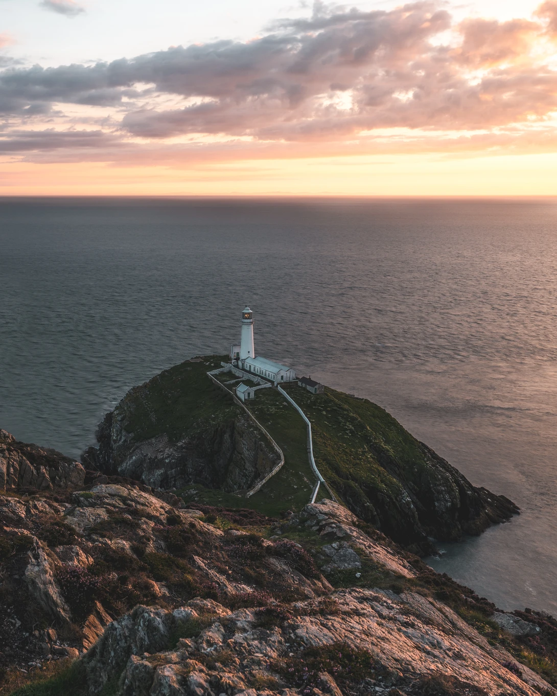 Lighthouse on a rocky headland at sunset with golden clouds and calm ocean waters.