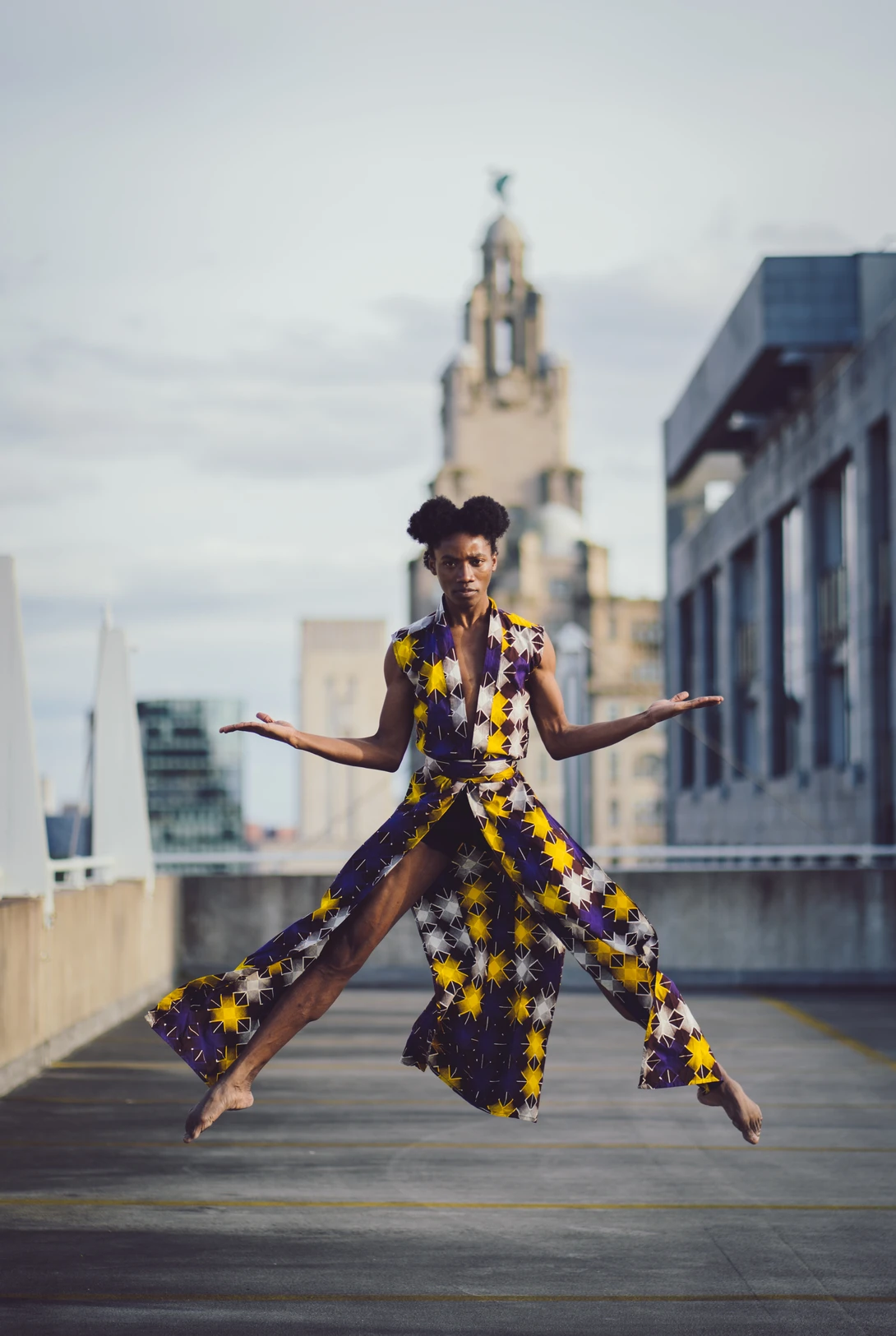 Dancer in yellow and purple patterned outfit mid-leap on an urban rooftop with a cathedral visible in the background.