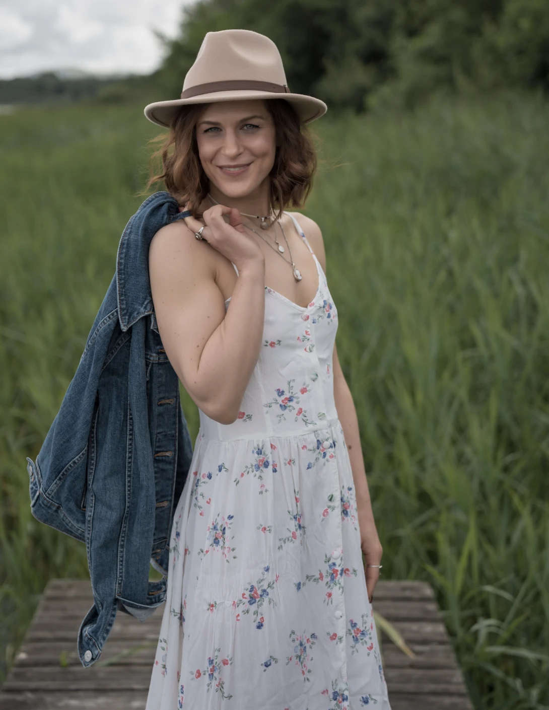 Woman in floral dress and denim jacket posing on wooden boardwalk in green field.