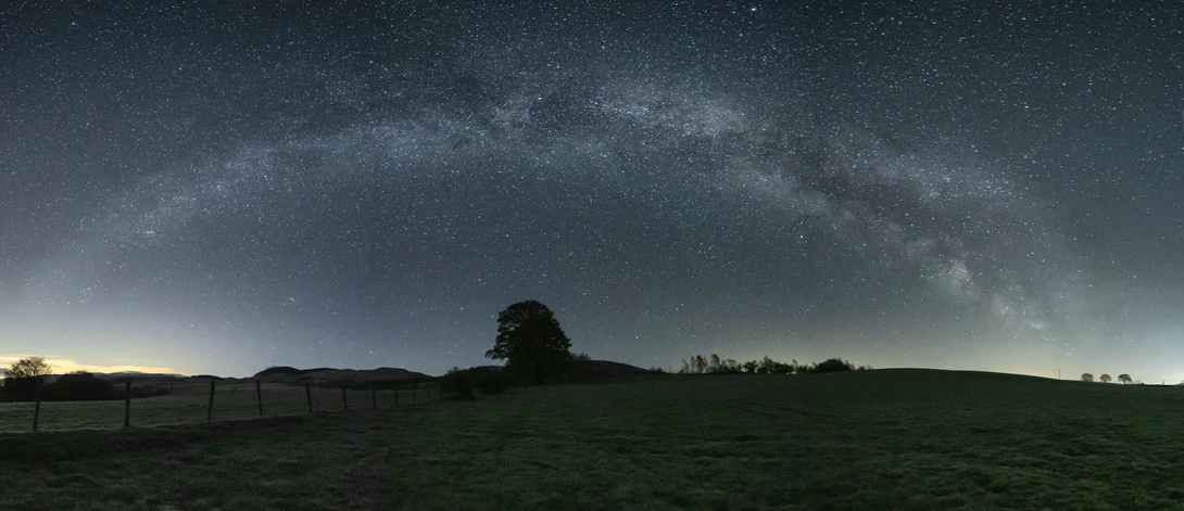 Milky Way galaxy stretching across night sky above rolling rural landscape with silhouetted trees and fields.