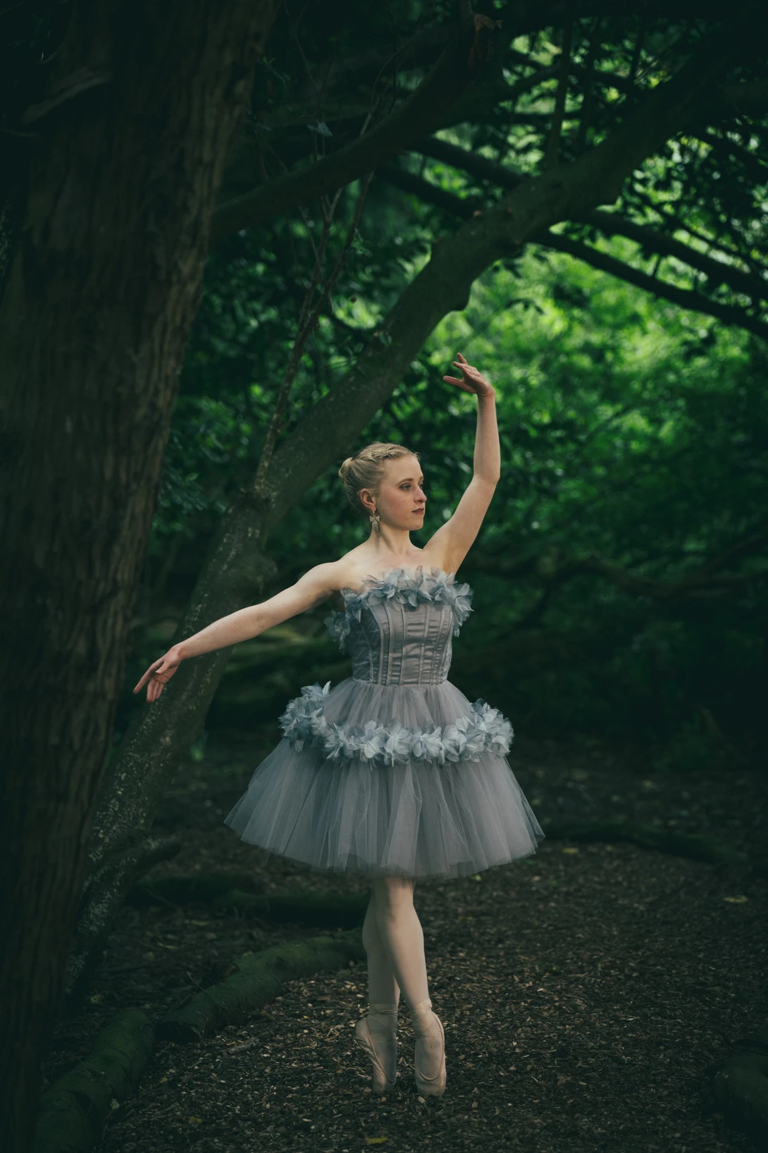 Ballet dancer in pointe shoes performing an arabesque pose on a forest path surrounded by dense green foliage.