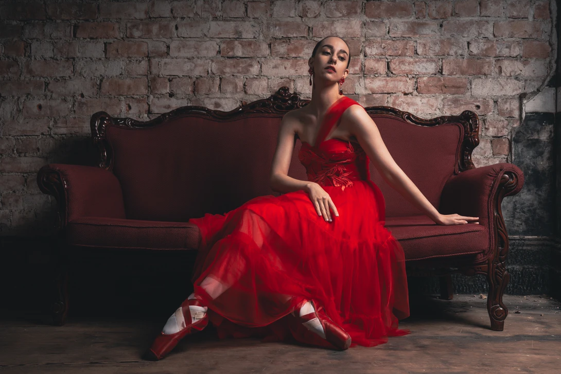 Woman in red ball gown seated on burgundy velvet sofa against exposed brick wall.