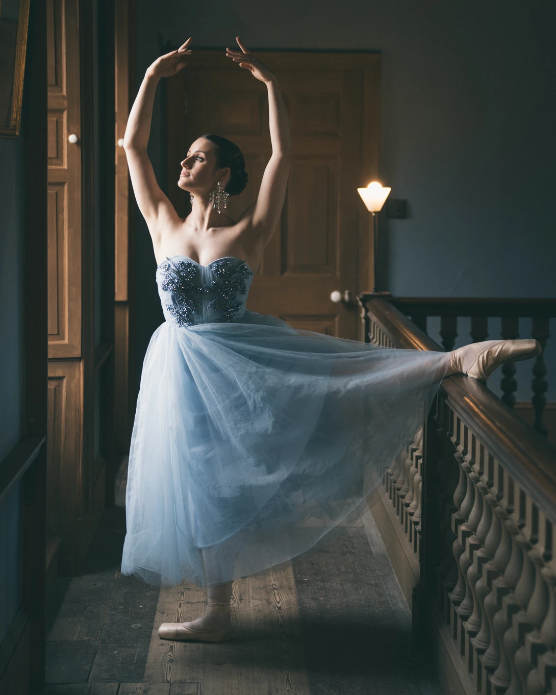 Ballet dancer in tulle skirt posed at stair railing with arms raised overhead.